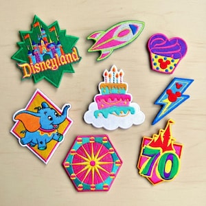 Puede incluir: Una colección de parches bordados coloridos con varios diseños. Incluyen un castillo de Disneyland, un cohete, una magdalena, un pastel, un rayo, un elefante, una noria y un diseño "70".