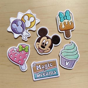 以下が含まれることがあります： ディズニーをテーマにした刺繍パッチのコレクション。ミッキーマウス、アイスクリーム、ハート型の棒付きキャンディー、そして「Magic & Dreams」と書かれたサインが含まれています。パッチは紫、黄色、ピンクなど、さまざまな色があります。