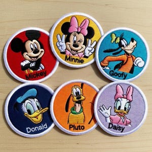 May include: Embroidered patches featuring Disney characters Mickey Mouse, Minnie Mouse, Goofy, Donald Duck, Pluto, and Daisy Duck. Each patch is round with a white border and a character's name below. The patches have vibrant colours.