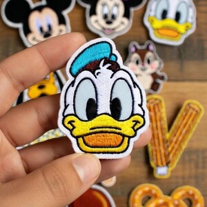 May include: A collection of embroidered patches featuring cartoon characters such as Mickey Mouse, Minnie Mouse, Donald Duck, and Chip and Dale. The patches are colourful and detailed, with a white border. Other patches include a pretzel and a fried chicken leg.