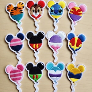May include: Twelve embroidered patches shaped like Mickey Mouse heads as balloons. Each patch features a different character or design, including Mickey, Stitch, and Chip. The patches have white borders and are on a light wood surface.