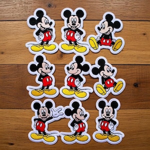 Puede incluir: Nueve parches de Mickey Mouse en varias poses. Cada parche presenta a Mickey con su atuendo clásico: pantalones cortos negros, pantalones rojos, zapatos amarillos y guantes blancos. Los parches están delineados en blanco y están sobre una superficie de madera.
