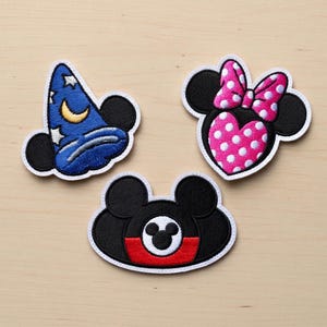 Puede incluir: Tres parches bordados con diseños de personajes de dibujos animados icónicos. Un parche representa un sombrero de mago azul con estrellas y una luna creciente. Otro muestra un corazón rosa con lunares blancos y un lazo. El tercer parche tiene un diseño rojo y negro.