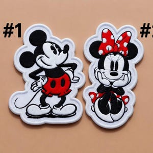 May include: Two embroidered patches featuring cartoon characters. One patch depicts Mickey Mouse in black, white, and red, with the number "#1" above it. The second patch shows Minnie Mouse in black, white, and red, with a red polka dot bow and the number "#2" above it.