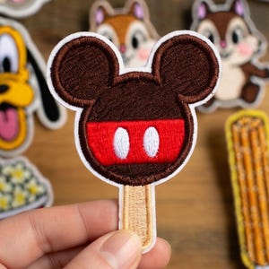 May include: Embroidered patch of a Mickey Mouse ice cream bar. The patch has a brown head with red and white details, and a beige stick. Other character patches are in the background.