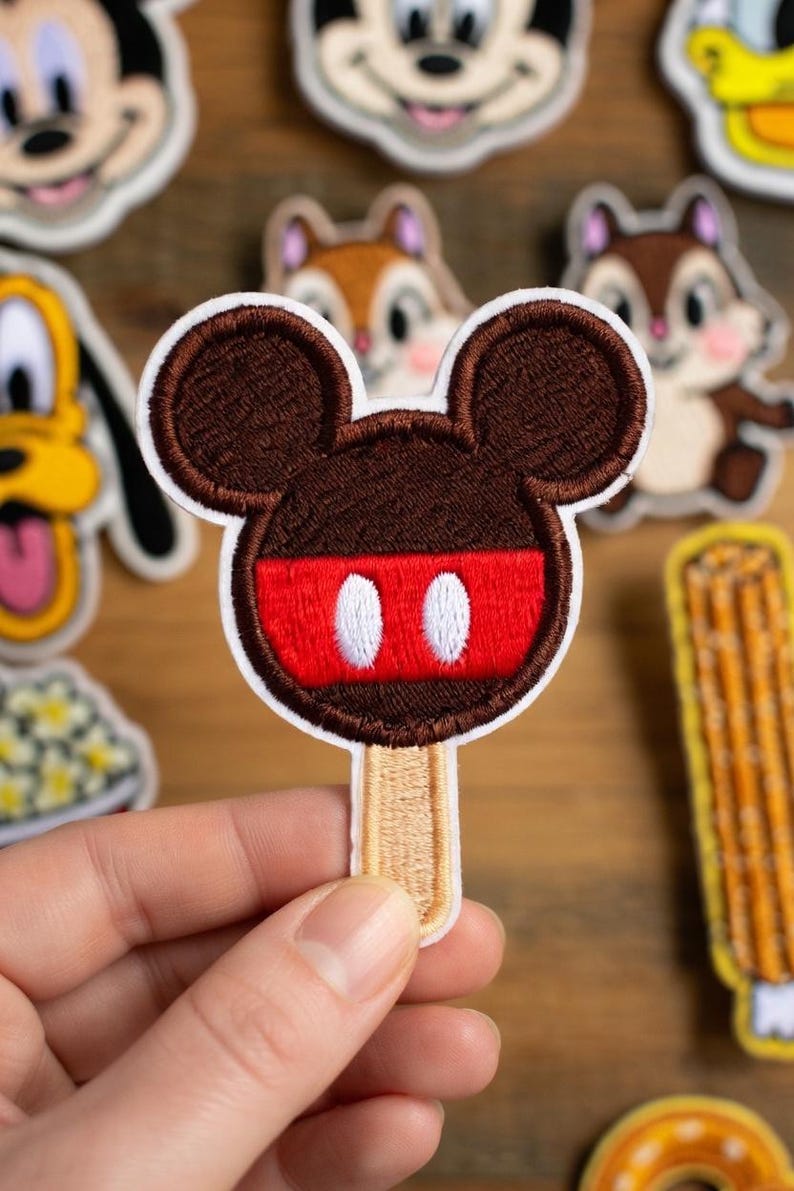 May include: Embroidered patch of a Mickey Mouse ice cream bar. The patch has a brown head with red and white details, and a beige stick. Other character patches are in the background.