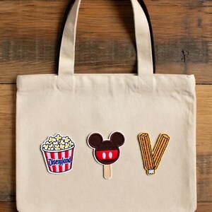 May include: Canvas tote bag in cream with black handles, featuring three embroidered patches: a popcorn bucket with "Disneyland" text, a Mickey Mouse ice cream, and a churro. The bag is made of canvas.