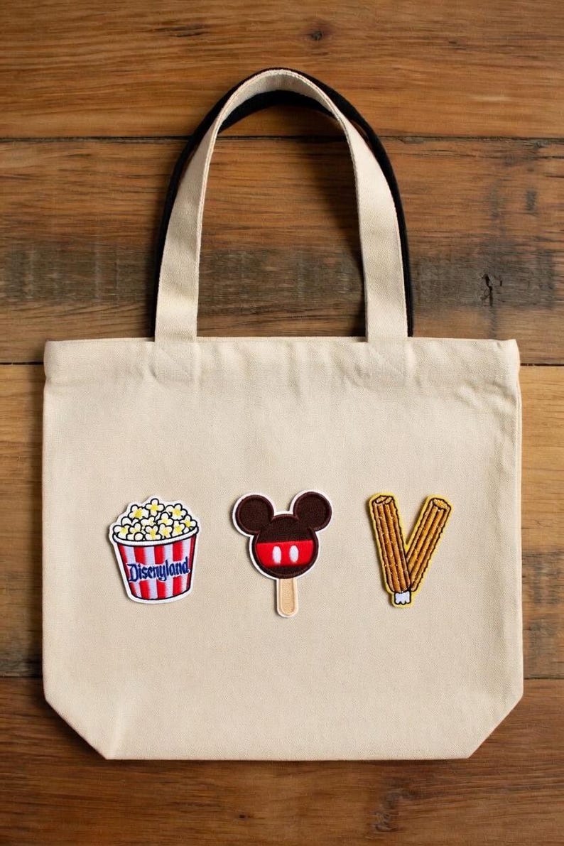 May include: Canvas tote bag in cream with black handles, featuring three embroidered patches: a popcorn bucket with "Disneyland" text, a Mickey Mouse ice cream, and a churro. The bag is made of canvas.