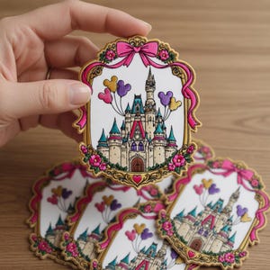 May include: A collection of decorative patches featuring a fairytale castle with colorful turrets, a pink bow, and Mickey Mouse balloon accents. The patches have a gold border with pink flowers and ribbons. The patches are held by a hand.