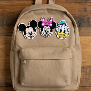 May include: A tan-coloured backpack featuring three character patches: Mickey Mouse, Minnie Mouse, and Donald Duck. The backpack has a front pocket with a zip and adjustable straps. The patches are colourful and add a playful touch to the bag.