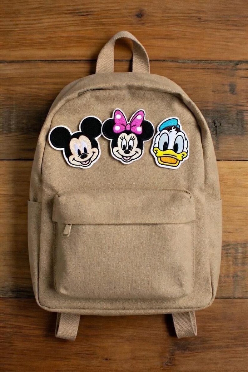 May include: A tan-coloured backpack featuring three character patches: Mickey Mouse, Minnie Mouse, and Donald Duck. The backpack has a front pocket with a zip and adjustable straps. The patches are colourful and add a playful touch to the bag.