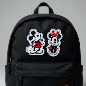 May include: Black backpack with two embroidered patches featuring Mickey Mouse and Minnie Mouse. Mickey is in a red shorts and Minnie is wearing a red polka dot bow. The patches are outlined in white.