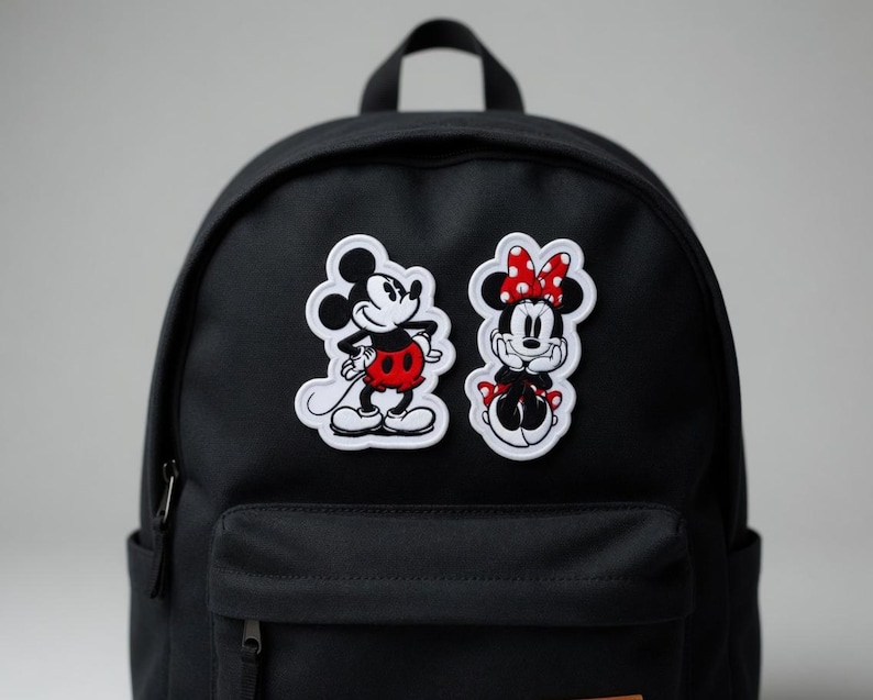 May include: Black backpack with two embroidered patches featuring Mickey Mouse and Minnie Mouse. Mickey is in a red shorts and Minnie is wearing a red polka dot bow. The patches are outlined in white.