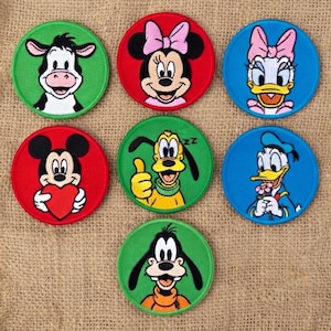 May include: Seven circular embroidered patches featuring cartoon characters. The patches have green, red, or blue backgrounds. Characters include a cow, Minnie Mouse, Daisy Duck, Mickey Mouse, Pluto, and Goofy. Each patch is circular.
