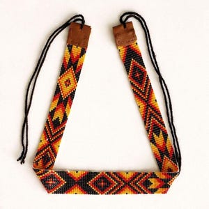 May include: A beaded necklace featuring a geometric pattern in black, red, orange, and yellow. The band is connected to brown leather tabs at the top, which are attached to thin black cords for tying. The design consists of repeating diamond shapes.