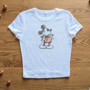 May include: White t-shirt featuring a hand-drawn style illustration of Mickey Mouse. The cartoon character is depicted in black, red, and white, with his hands on his hips. The shirt has a crew neck and short sleeves.