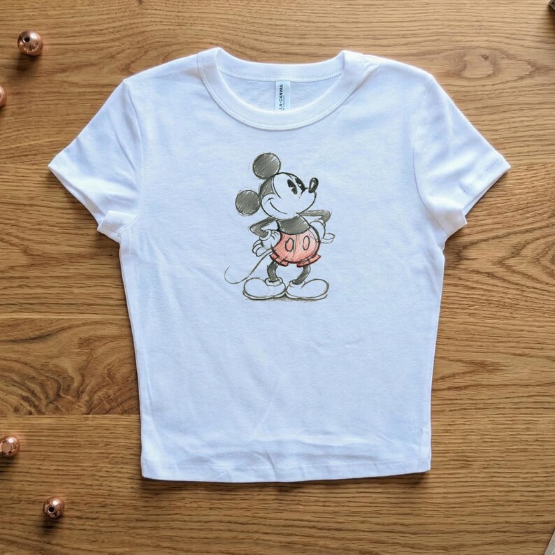 May include: White t-shirt featuring a hand-drawn style illustration of Mickey Mouse. The cartoon character is depicted in black, red, and white, with his hands on his hips. The shirt has a crew neck and short sleeves.