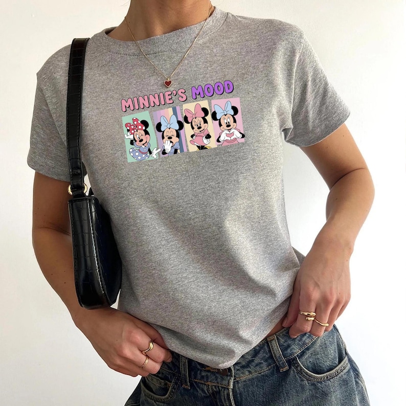 May include: A gray t-shirt featuring the text "MINNIE'S MOOD" above a graphic of Minnie Mouse in four different poses. The graphic has a pastel color scheme. The shirt is short-sleeved.