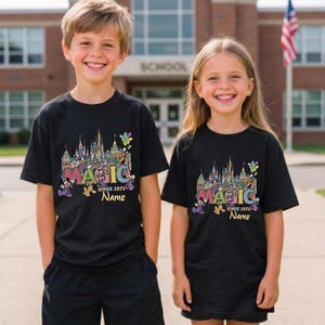May include: Two children wearing black t-shirts with a colorful graphic of a castle, balloons, and the word "MAGIC". The shirts also feature the text "SINCE 1971" and a space for a name.