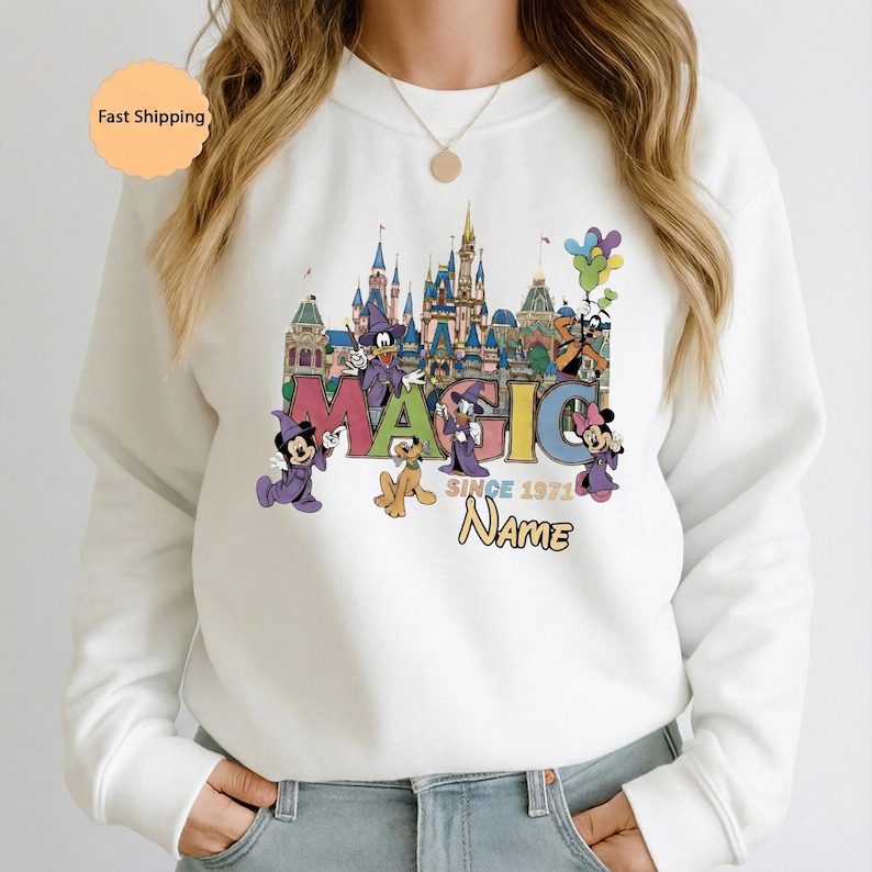 May include: White sweatshirt featuring a colorful graphic of a castle, Disney characters, and the word "MAGIC" in large letters. The design includes the text "SINCE 1971" and a space for a name. The sweatshirt has long sleeves.