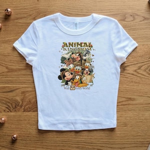 K&ouml;nnte beinhalten: Wei&szlig;es T-Shirt mit einer farbenfrohen Grafik von Disney-Figuren, darunter Micky Maus, Donald Duck und Goofy. Der Text "ANIMAL KINGDOM" und "Walt Disney World" ist ebenfalls auf dem T-Shirt abgebildet.