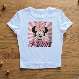 May include: White t-shirt featuring a graphic of Minnie Mouse with a pink bow and the name "Minnie" in pink script. The design has a retro, distressed look with pink and white rays. The shirt is made of a soft material.