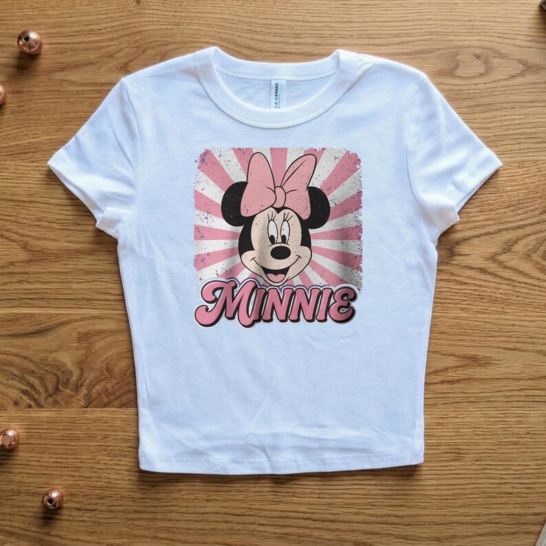 May include: White t-shirt featuring a graphic of Minnie Mouse with a pink bow and the name "Minnie" in pink script. The design has a retro, distressed look with pink and white rays. The shirt is made of a soft material.