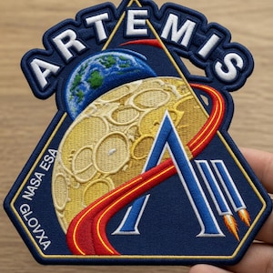 May include: Embroidered patch featuring the word "ARTEMIS" in white letters above a blue and gold design. The design includes a blue Earth, a yellow moon, and a red and yellow curved path. The patch also includes the text "NASA ESA GLOVXA".