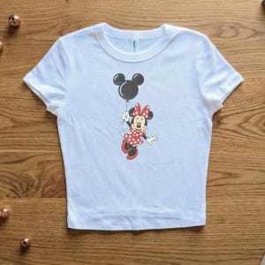 May include: White t-shirt featuring a graphic of Minnie Mouse holding a black balloon. Minnie Mouse is wearing a red polka dot dress and bow. The shirt is made of a soft material.