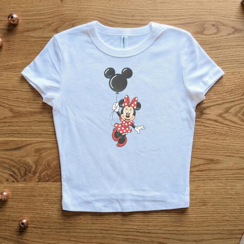 May include: White t-shirt featuring a graphic of Minnie Mouse holding a black balloon. Minnie Mouse is wearing a red polka dot dress and bow. The shirt is made of a soft material.
