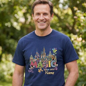 May include: Navy blue t-shirt featuring a colorful graphic of a castle, balloons, and cartoon characters. The word "MAGIC" is displayed prominently, along with "SINCE 1971" and a space for a name. The shirt is a classic crew neck style.