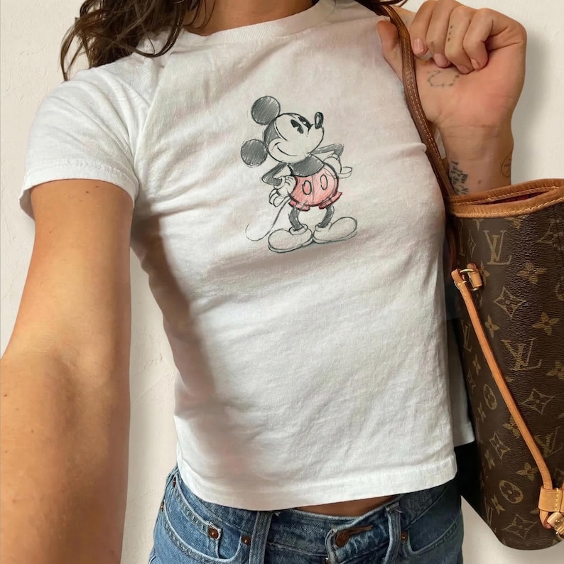 May include: White t-shirt featuring a hand-drawn sketch of Mickey Mouse. The cartoon character is depicted in black and white with a pink skirt. The shirt is paired with blue jeans and a brown handbag.