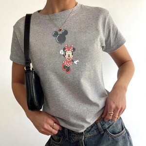 May include: A gray t-shirt featuring a cartoon Minnie Mouse design. Minnie Mouse is wearing a red polka dot dress and bow. The shirt has short sleeves and a crew neck. The image also shows a black purse and jewelry.