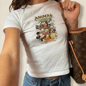 K&ouml;nnte beinhalten: Wei&szlig;es T-Shirt mit einer bunten Grafik von Disney-Figuren, darunter Micky Maus, Donald Duck und Goofy. Das Shirt tr&auml;gt den Text "ANIMAL KINGDOM" und "Walt Disney World". Das Shirt wird mit einer blauen Jeans kombiniert.