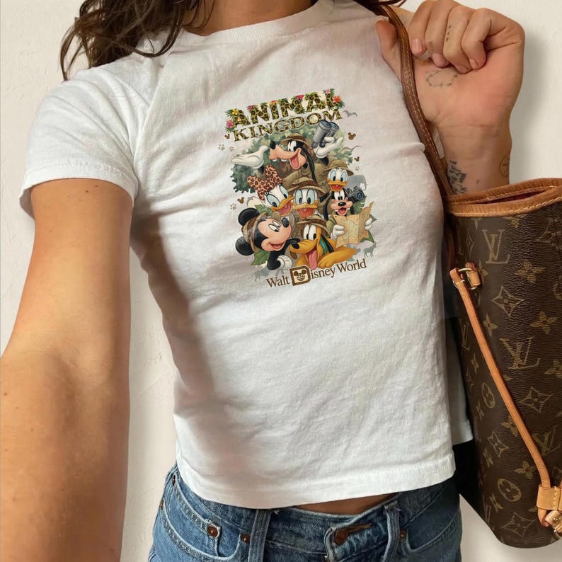 K&ouml;nnte beinhalten: Wei&szlig;es T-Shirt mit einer bunten Grafik von Disney-Figuren, darunter Micky Maus, Donald Duck und Goofy. Das Shirt tr&auml;gt den Text "ANIMAL KINGDOM" und "Walt Disney World". Das Shirt wird mit einer blauen Jeans kombiniert.