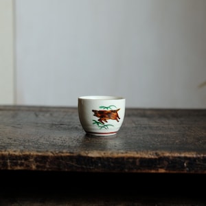 May include: A small, off-white ceramic cup with a brown and green illustration of a running animal. The cup has a red band at the base and a cream-colored rim. The cup is set on a dark wooden surface.
