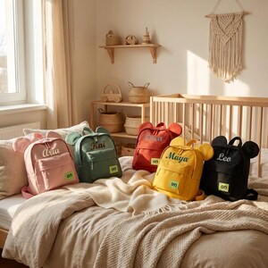 Peut inclure: Cinq petits sacs à dos avec des oreilles arrondies sur le dessus, chacun portant un prénom brodé en écriture cursive et un petit écusson carré vert avec le texte 'on'. Les sacs sont rose, vert sauge, rouge, jaune et noir, posés sur un lit.