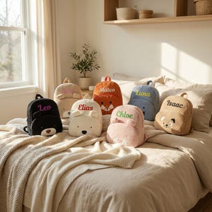Puede incluir: Una colección de siete mochilas pequeñas de peluche con temática animal en varios colores, incluyendo negro, beige, naranja, azul y rosa. Cada mochila presenta un nombre bordado único: Leo, Aria, Elias, Mateo, Luna, Chloe e Isaac.