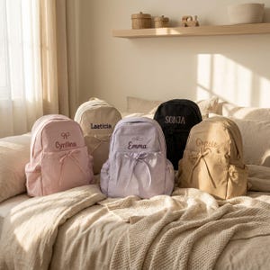Könnte beinhalten: Fünf Rucksäcke in verschiedenen Farben, darunter Hellrosa, Beige, Lavendel und Schwarz. Jeder Rucksack verfügt über eine Vordertasche mit Kordelzug und ist mit den gestickten Namen Cyntina, Laeticia, Emma, Sonja und Gracie personalisiert.