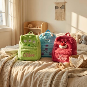 May include: Three plush backpacks featuring character designs: a lime green alien with three eyes labeled Chloe, a blue monster with horns labeled Felix, and a dark pink bear with a white muzzle labeled Leo. Each bag has a front zippered pocket.