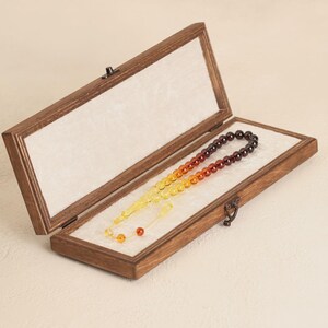 May include: A wooden box with a hinged lid, lined with white fabric, displays a prayer bead set. The beads transition in color from yellow to orange to brown. The box is open, revealing the beads.