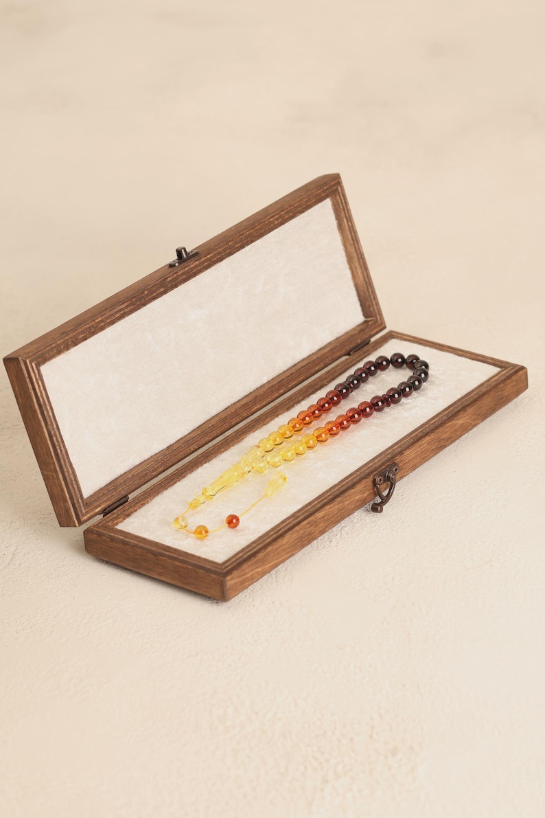 May include: A wooden box with a hinged lid, lined with white fabric, displays a prayer bead set. The beads transition in color from yellow to orange to brown. The box is open, revealing the beads.
