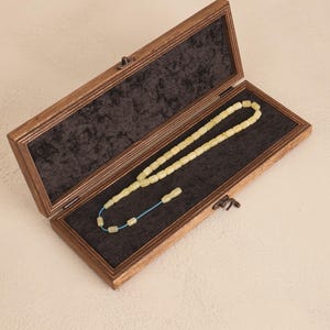 Pode incluir: Um conjunto de contas de ora&ccedil;&atilde;o de cor clara, exibido dentro de uma caixa de madeira aberta com um forro de veludo cinza escuro. As contas s&atilde;o conectadas por um fio azul claro. A caixa tem um fecho simples.