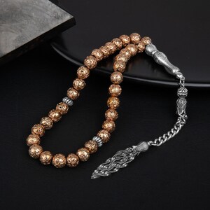 May include: A prayer bead strand featuring spherical, textured copper-colored beads interspersed with small silver-toned spacers. The strand terminates in a decorative silver-toned metal tassel component with a chain link and a textured leaf-like end.