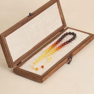 May include: A wooden box with a hinged lid, containing a prayer bead set. The beads transition in color from yellow to orange to dark brown. The box is lined with white fabric and has a metal clasp.