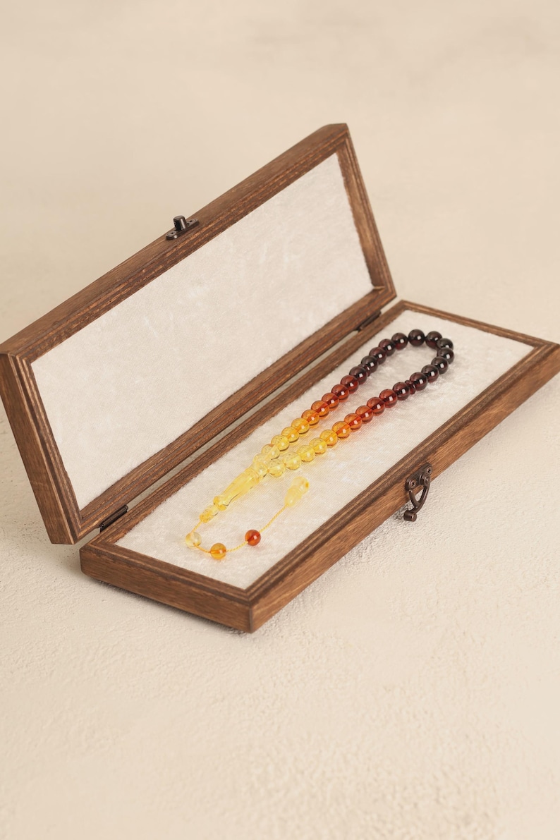 May include: A wooden box with a hinged lid, containing a prayer bead set. The beads transition in color from yellow to orange to dark brown. The box is lined with white fabric and has a metal clasp.