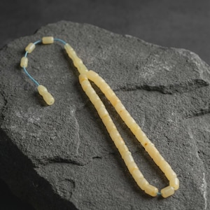 Pode incluir: Um colar de contas de ora&ccedil;&atilde;o amarelo claro com um fio azul, repousando sobre uma superf&iacute;cie de pedra cinza escuro. As contas s&atilde;o cil&iacute;ndricas e t&ecirc;m uma apar&ecirc;ncia ligeiramente transl&uacute;cida. O colar &eacute; um item religioso tradicional.