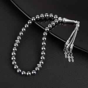 May include: A string of polished, dark gray spherical beads arranged in a loop. The strand features two decorative silver-toned metal spacers and terminates in a silver-toned metal tassel assembly with three dangling leaf-shaped charms.