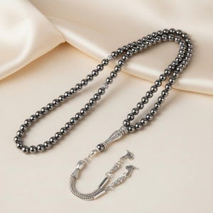 May include: A dark grey beaded rosary with a silver-coloured decorative pendant and chain. The beads are round and evenly spaced. The background is a light cream colour.