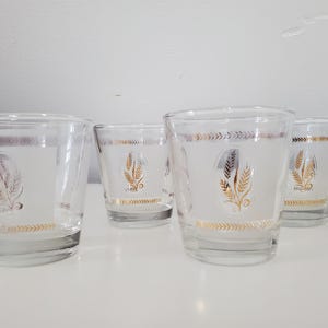 May include: Four clear glass tumblers with a frosted design. Each glass has a gold wheat stalk and decorative gold bands. The glasses are displayed on a white surface.
